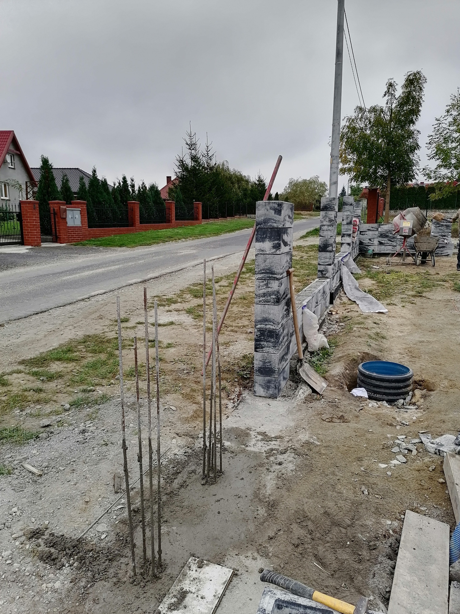 Budowa ogrodzenia z betonowych bloczków w Kunicach. Widoczne wystające pręty zbrojeniowe, zaprawa, narzędzia i częściowo wzniesiona konstrukcja.