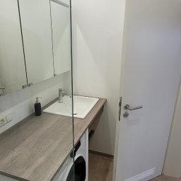 ARTEX - Nowoczesna łazienka z pralką zabudowaną pod blatem umywalki, minimalistyczne szafki z lustrami, szklana ścianka prysznicowa i beżowa podłoga.