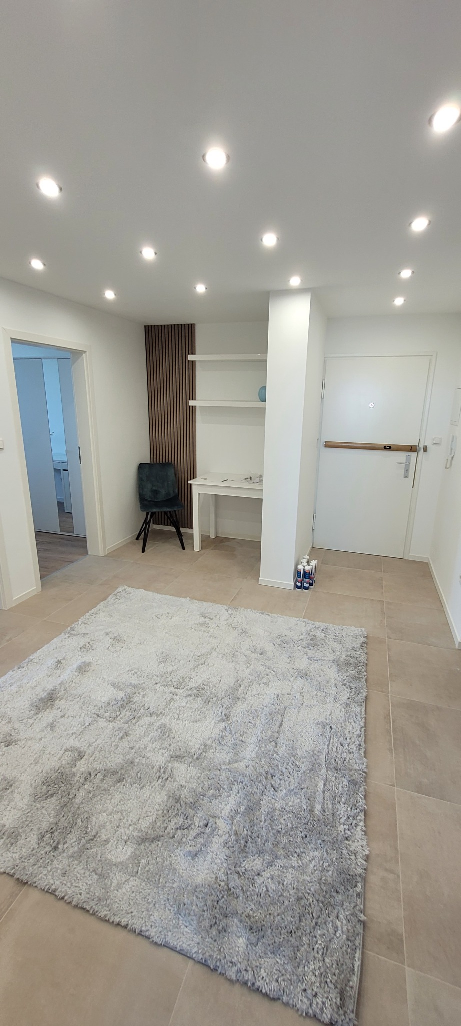 Jasne wnętrze po remoncie: białe ściany, beżowe płytki, szary dywan, punktowe oświetlenie sufitowe. Widoczne drzwi, krzesło i półki. Minimalistyczny design.