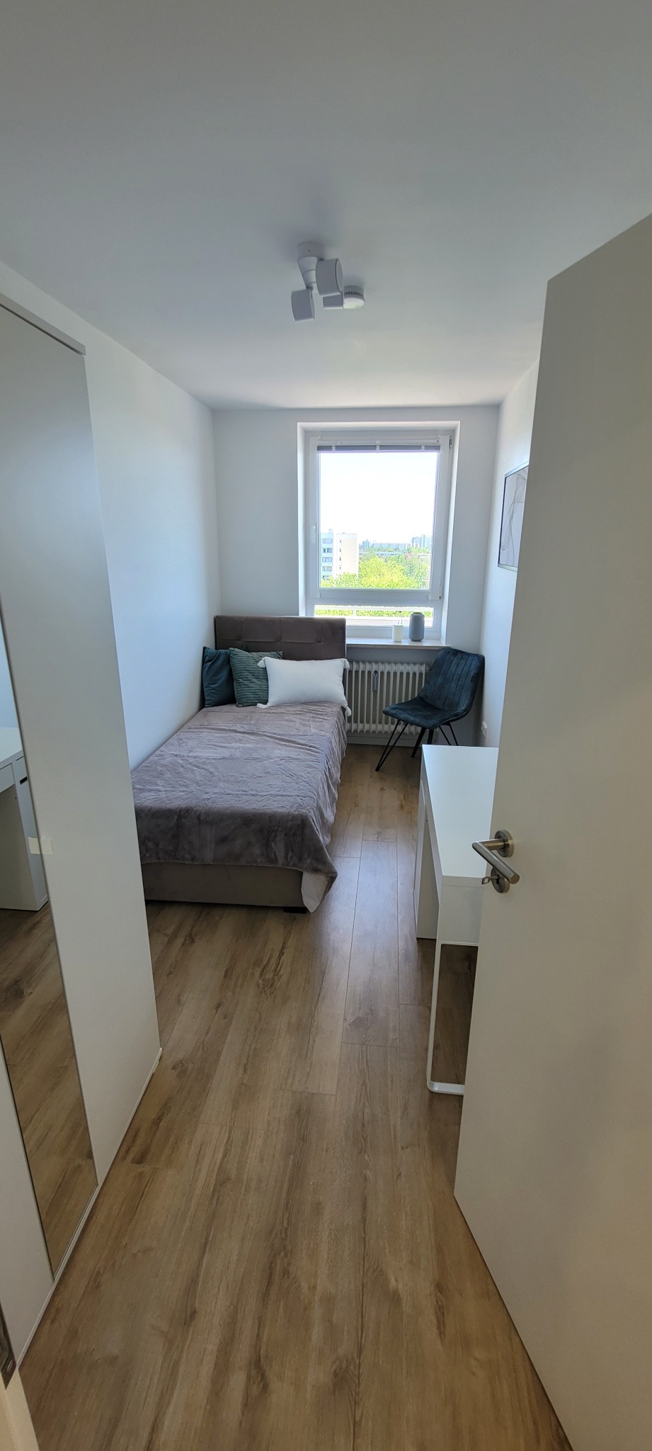 Jasne wnętrze pokoju z łóżkiem, biurkiem i szafą. Drewniana podłoga i białe ściany tworzą nowoczesny wygląd. Widok z perspektywy wejścia.