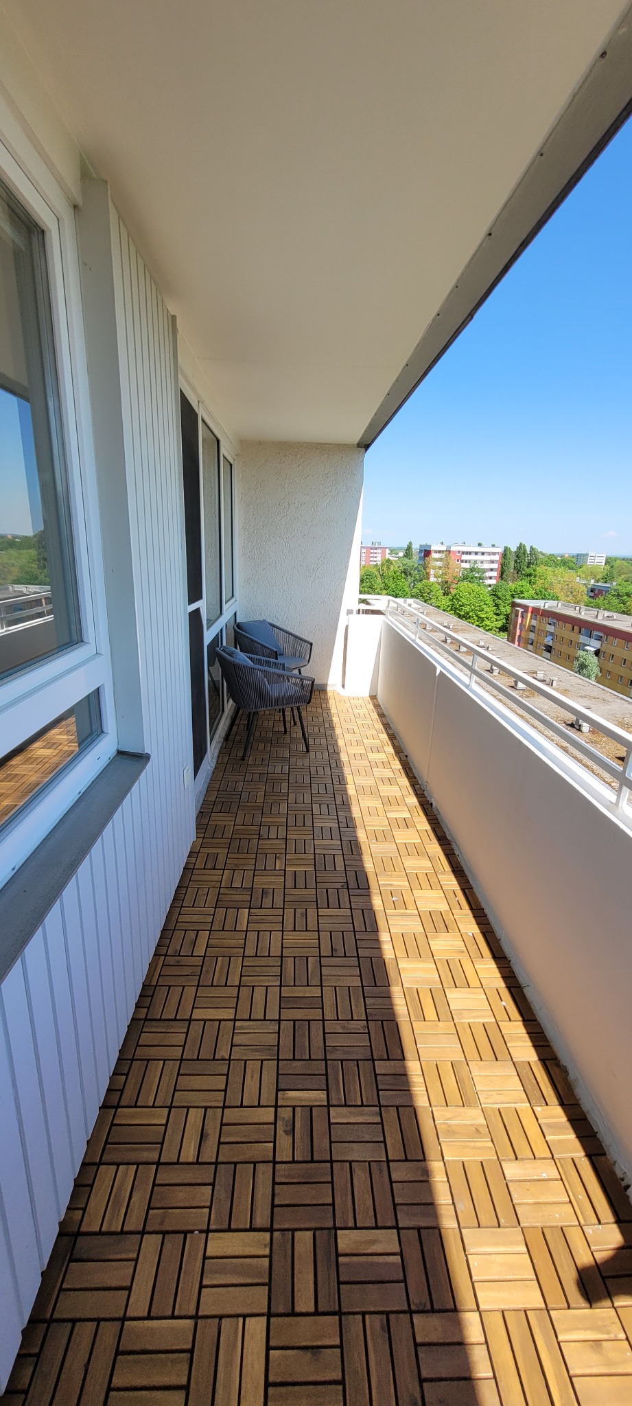 Jasny balkon z drewnianą podłogą w kwadraty, dwa krzesła z plecionki i widok na zieleń miejską w słoneczny dzień. Minimalistyczny design.