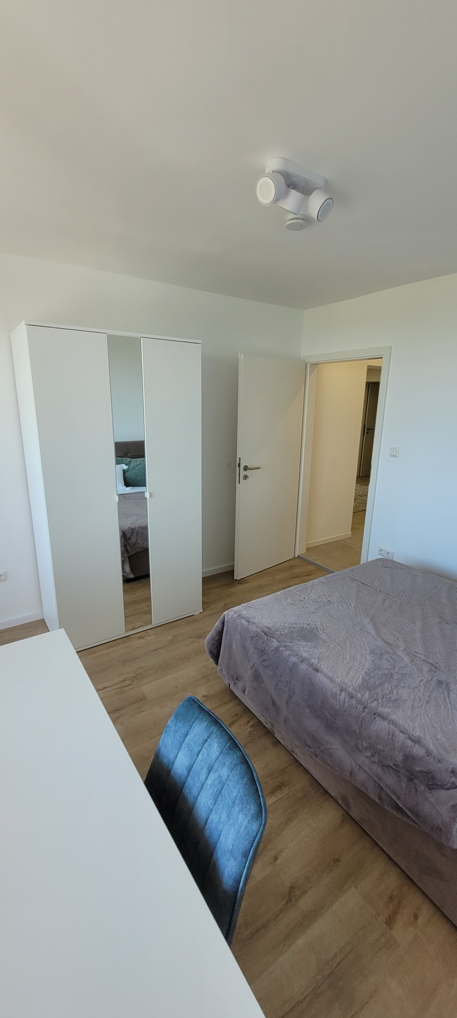 Jasne wnętrze sypialni z łóżkiem, szafą z lustrem, biurkiem i krzesłem na drewnianej podłodze. Minimalistyczny design z białymi ścianami i nowoczesnym oświetleniem.