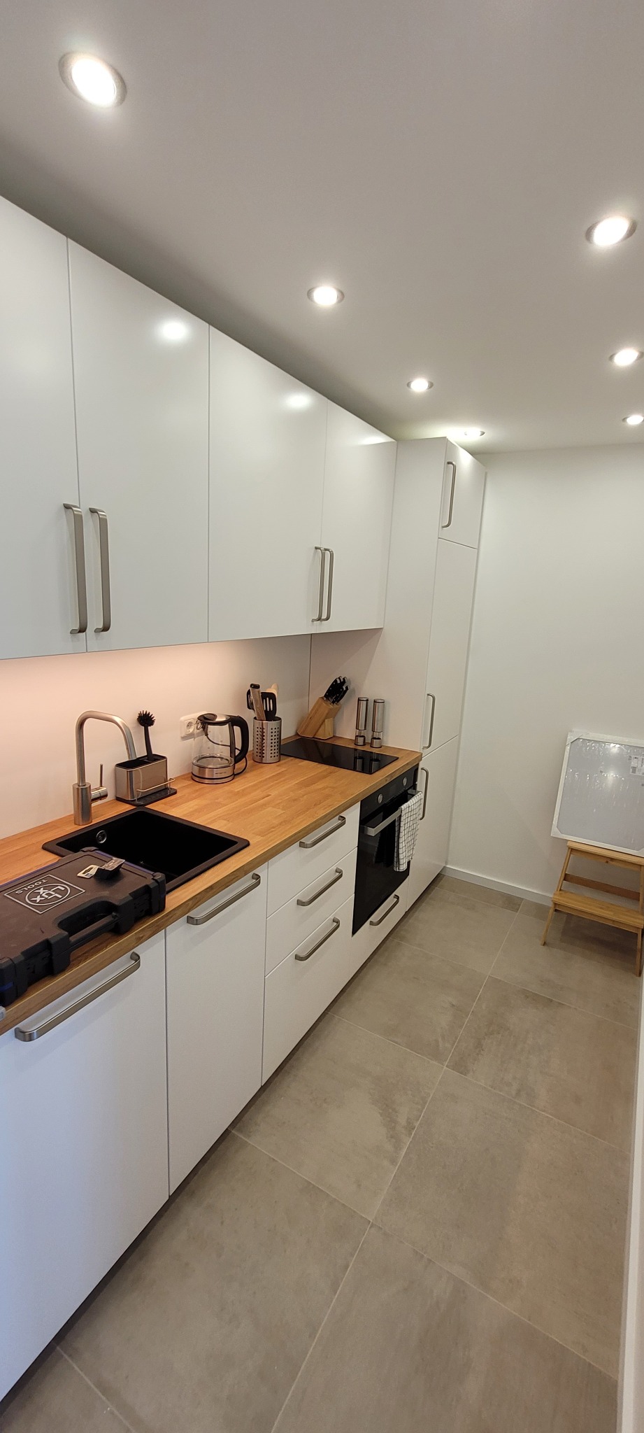 Biała kuchnia z drewnianym blatem, czarnym zlewem i płytą indukcyjną. Na blacie narzędzia, oświetlenie punktowe sufitowe. Minimalistyczny styl.