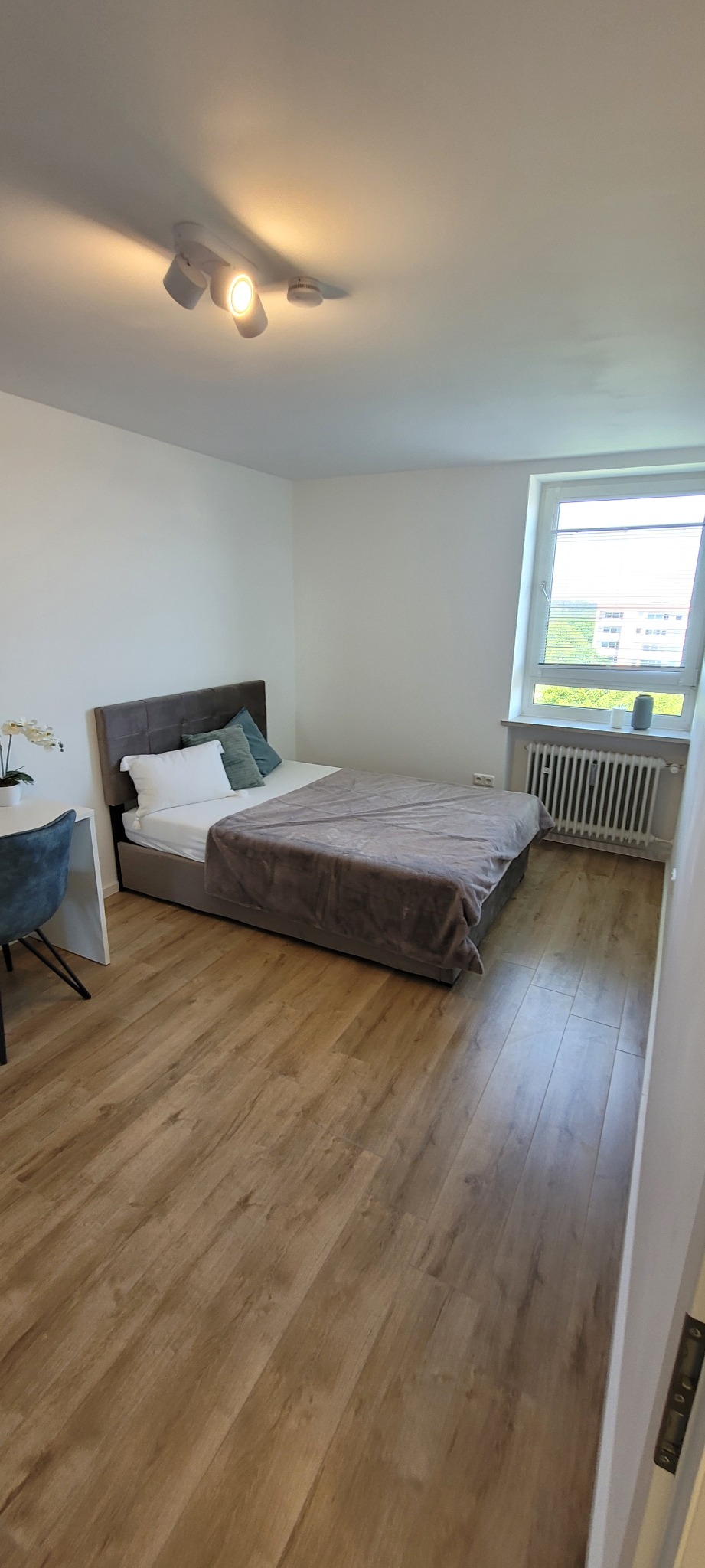 Jasny pokój w Warszawie po wykończeniu: drewniana podłoga, szare łóżko, białe ściany i sufit z oświetleniem punktowym. Okno z widokiem na zieleń.