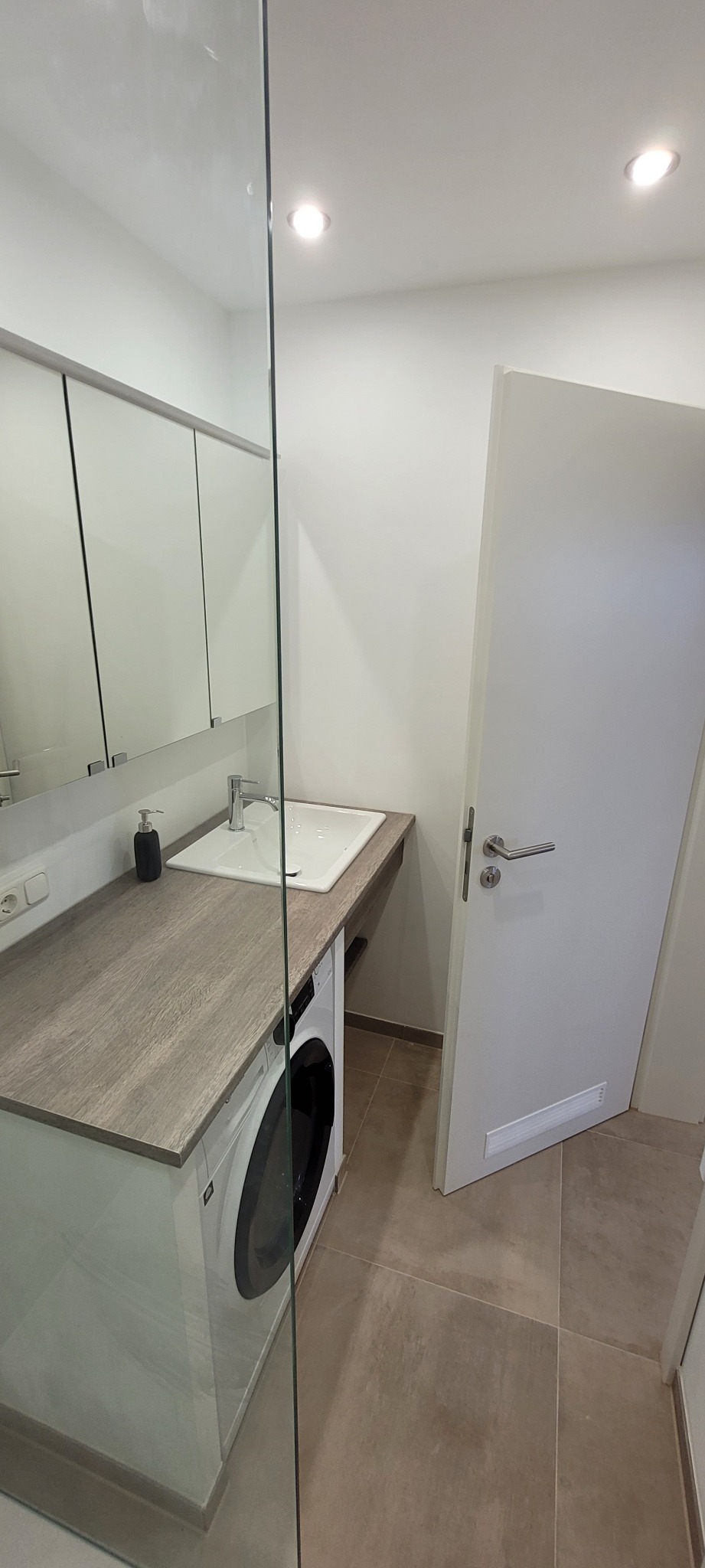 Nowoczesna łazienka z pralką zabudowaną pod blatem umywalki, minimalistyczne szafki z lustrami, szklana ścianka prysznicowa i beżowa podłoga.