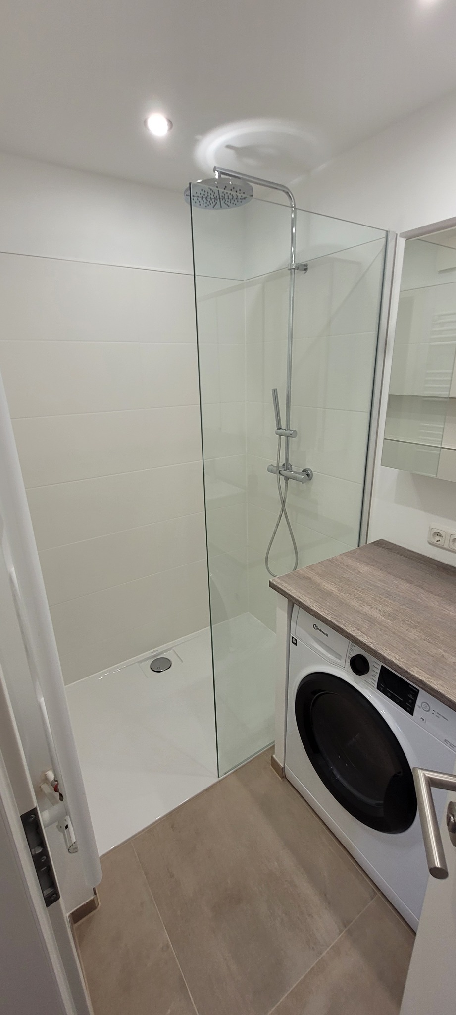 Nowoczesna łazienka z kabiną prysznicową typu walk-in, białe płytki i pralka pod blatem. Minimalistyczny design z chromowanymi detalami.