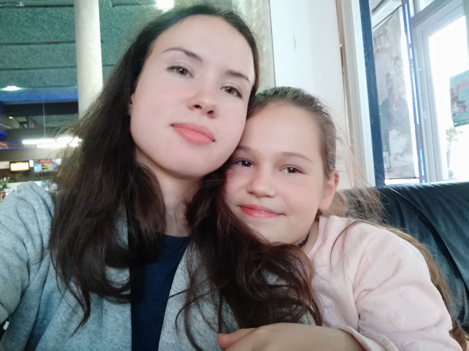 Portret uśmiechniętej nastolatki obejmującej młodszą dziewczynkę na tle wnętrza z oknem. Zdjęcie wykonane z bliska, w naturalnym oświetleniu.