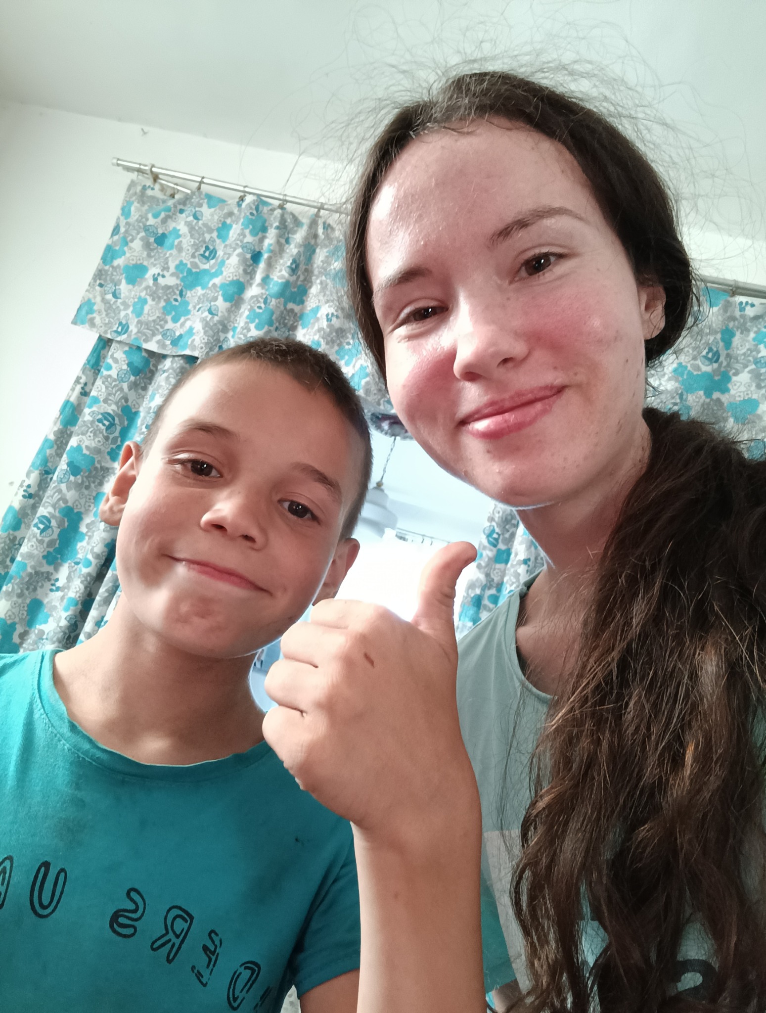 Uśmiechnięta niania z chłopcem, kciuk w górę, w tle zasłony w kwiaty. Zdjęcie z bliska, domowe otoczenie, pozytywna atmosfera.