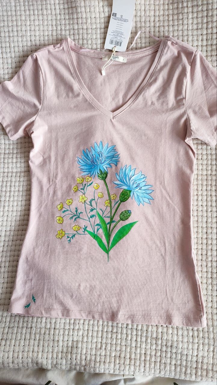 Różowa koszulka z ręcznie malowanymi błękitnymi chabrami i żółtymi kwiatkami, leżąca na kremowej narzucie. Widoczna metka.