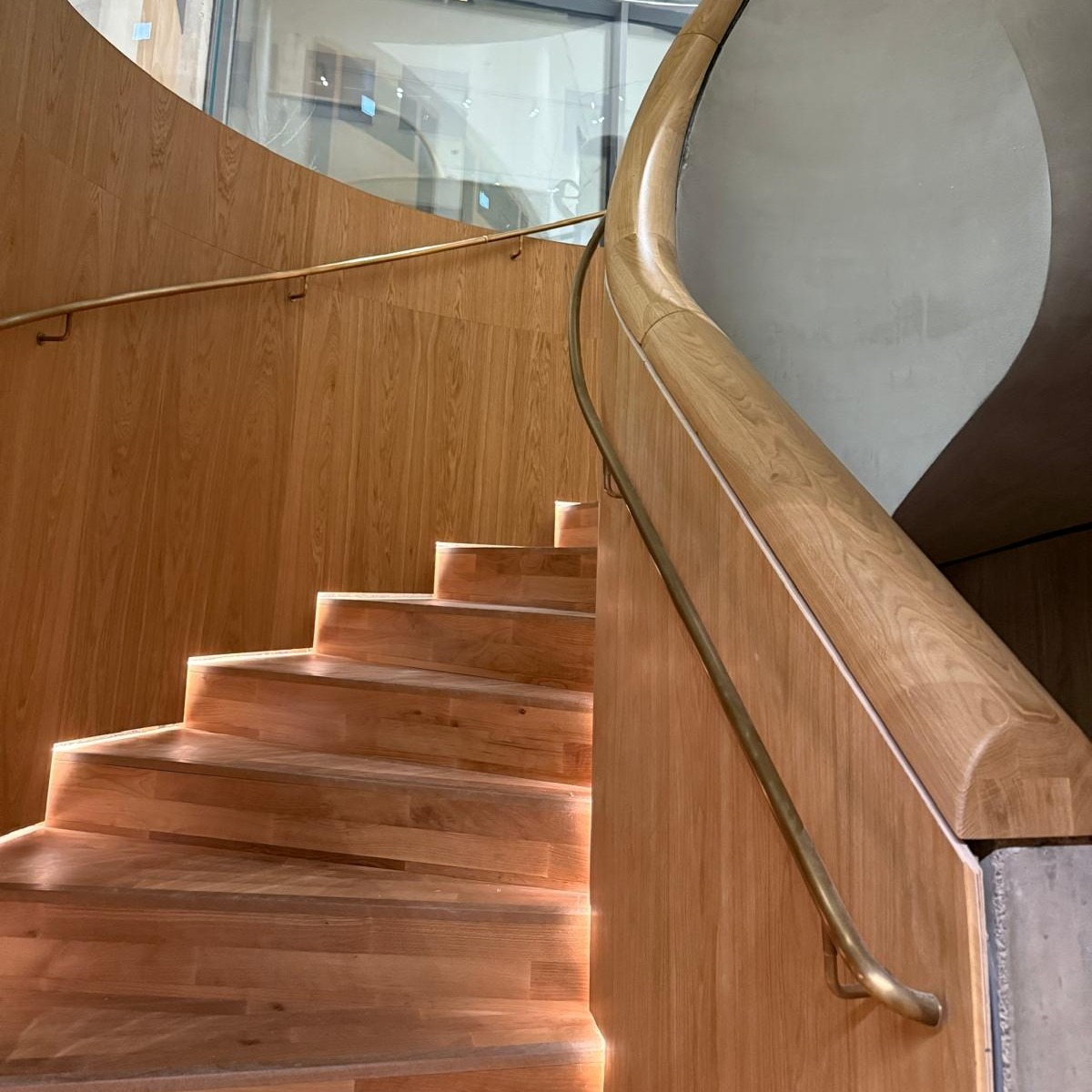 Drewniane schody spiralne z oświetleniem LED pod stopniami i mosiężną poręczą. Widok z dołu, podkreślający zakrzywiony kształt i detale wykończenia.
