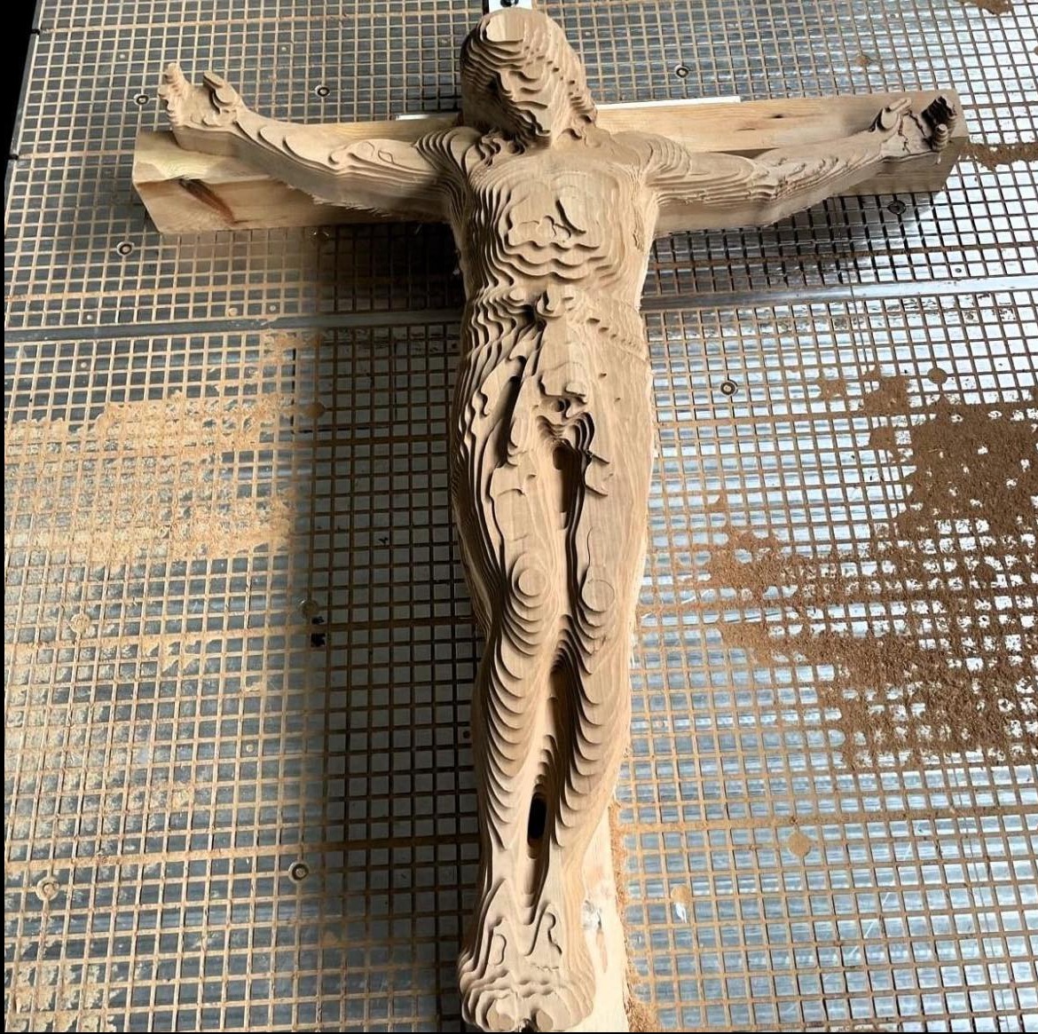 Drewniana rzeźba Jezusa na krzyżu, wykonana warstwowo, leżąca na metalowej siatce. Widoczne ślady obróbki, jasne drewno.