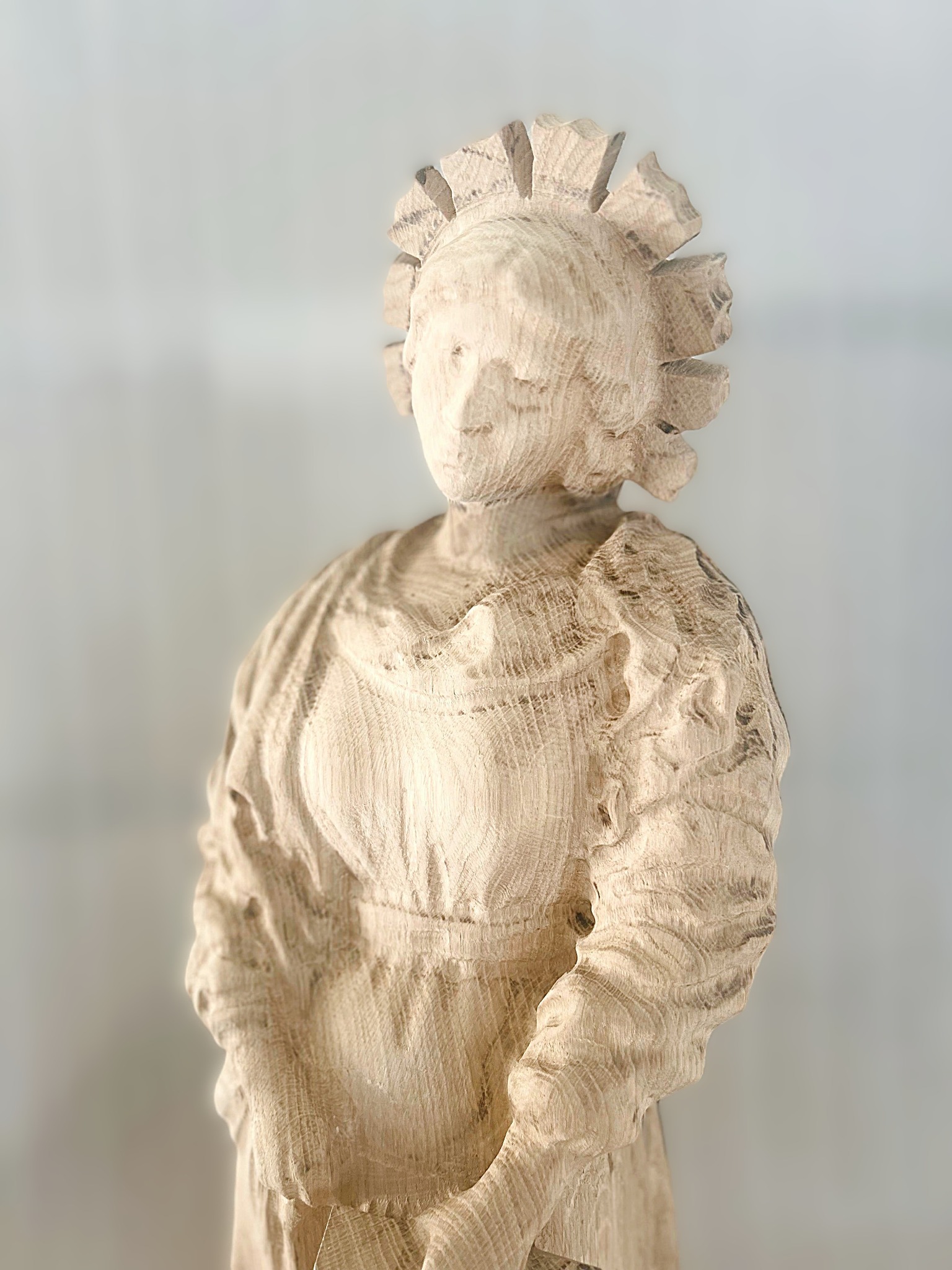 Drewniana rzeźba kobiety w tradycyjnym stroju, z widocznymi śladami obróbki dłutem, na rozmytym, jasnym tle. Artystyczne detale i faktura drewna.