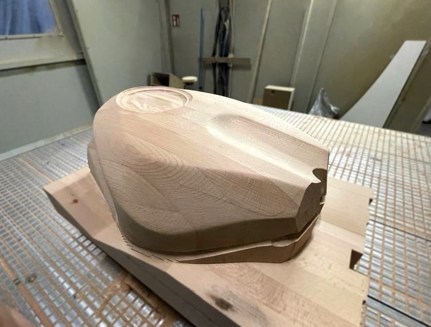 Drewniany model zbiornika paliwa motocykla, precyzyjnie wyfrezowany na stole roboczym z widocznymi śladami obróbki. Jasne drewno, neutralne tło warsztatu.