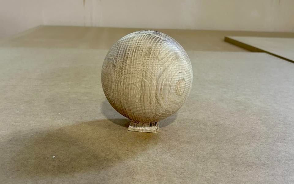 Drewniana kula z widocznymi słojami, osadzona na małej podstawce, na neutralnym tle. Precyzyjna obróbka drewna, ukazująca strukturę materiału.