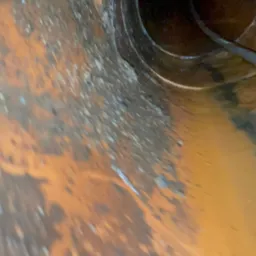 Wnętrze brudnej, pomarańczowej rury kanalizacyjnej z widoczną pajęczyną i osadami. Nietypowy kąt ujęcia podkreśla głębię i stan zaniedbania.