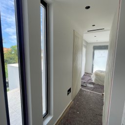 UBS - Wnętrze w trakcie wykończenia: białe ściany, zabezpieczona podłoga i meble folią malarską. Widoczne okna z czarnymi ramami i fragment zieleni za oknem.