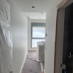 UBS - Biały korytarz w trakcie malowania: świeżo pomalowane ściany, zabezpieczone okno folią, podłoga przykryta materiałem ochronnym, widoczne wejście do łazienki.