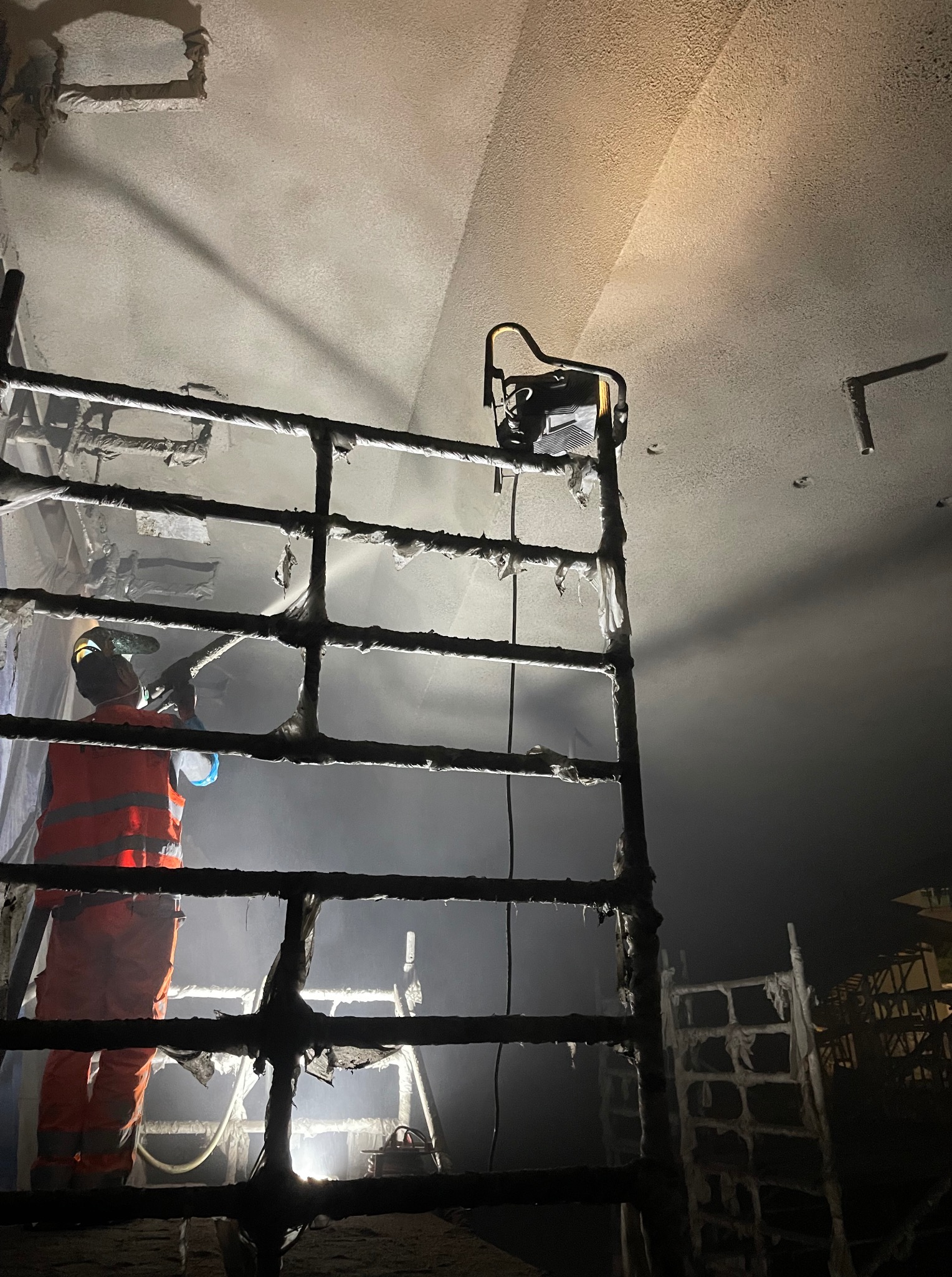 Pracownik w kombinezonie spryskuje sufit z rusztowania, oświetlony lampą. Widoczne zabezpieczenia i narzędzia pracy. Nocna sceneria, detale wykończenia wnętrz.