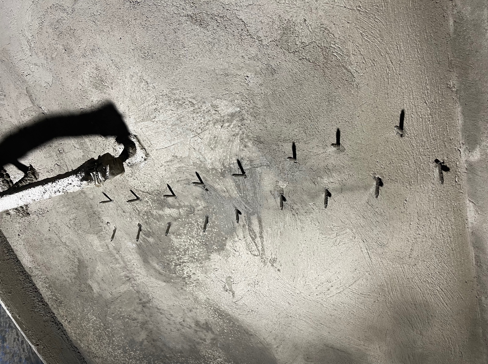 Świeżo wylany beton z wystającymi prętami zbrojeniowymi, widoczne ślady po szalunku i cień węża do betonu. Surowy stan w trakcie prac budowlanych.