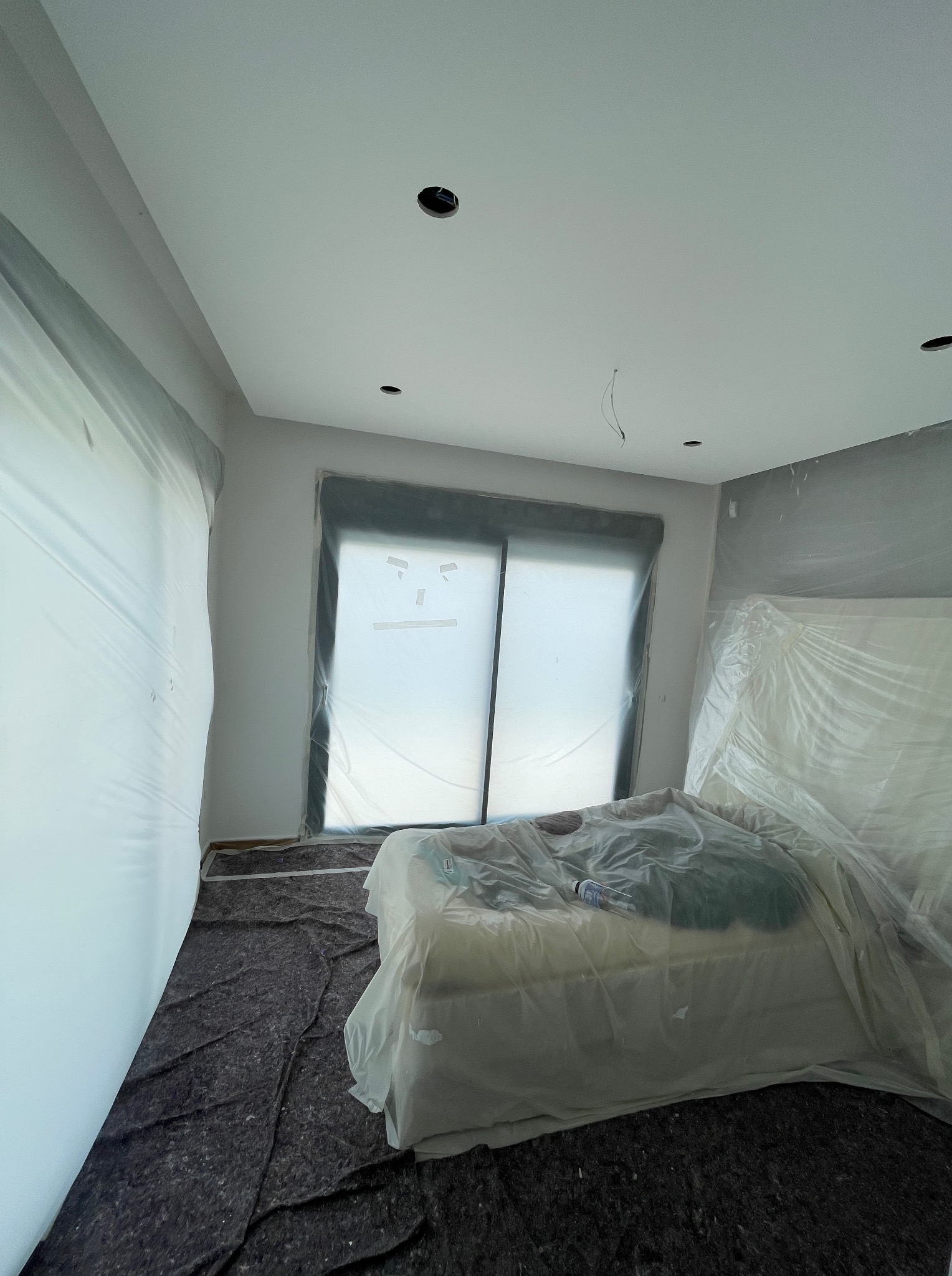 Pokój zabezpieczony folią malarską przed malowaniem: okna, ściany i meble osłonięte. Widoczne przygotowania do malowania ścian na biało.