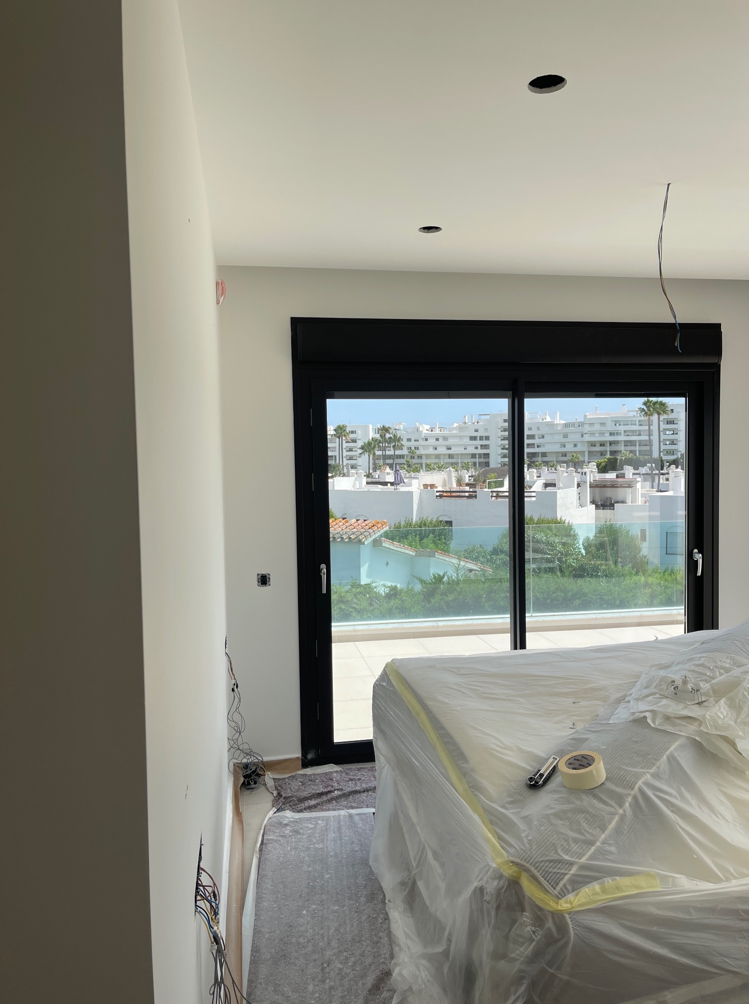 Wnętrze w trakcie prac wykończeniowych: ściany przygotowane do malowania, okno z czarną ramą, meble zabezpieczone folią. Widoczne kable i otwory w suficie.