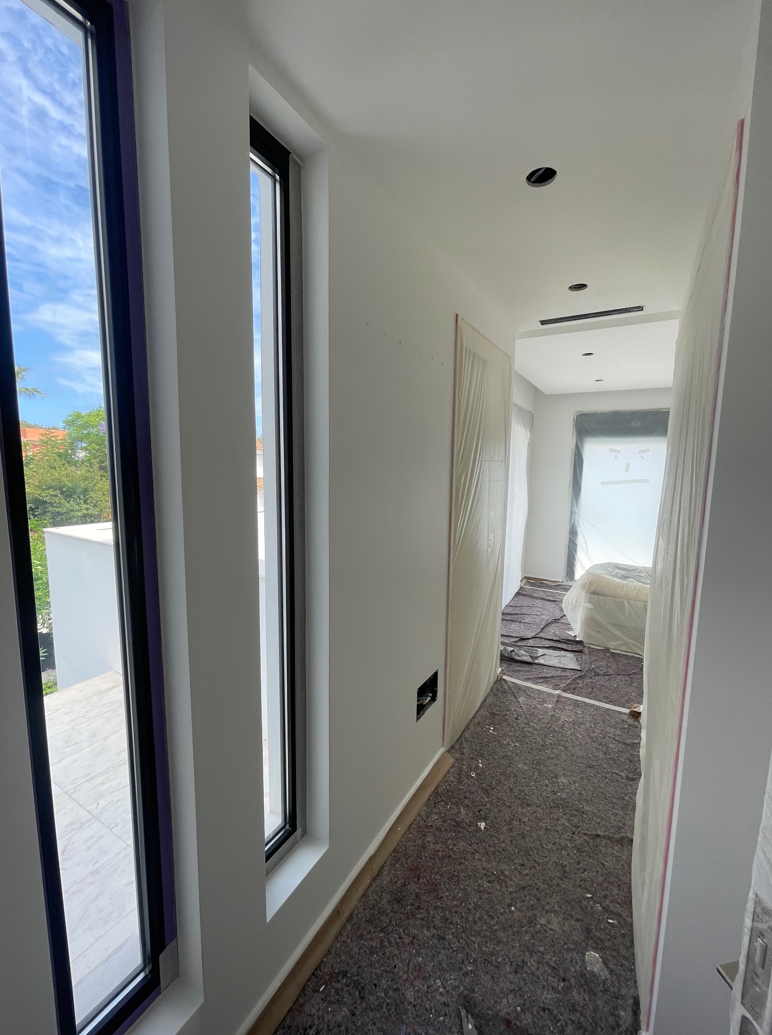 Wnętrze w trakcie wykończenia: białe ściany, zabezpieczona podłoga i meble folią malarską. Widoczne okna z czarnymi ramami i fragment zieleni za oknem.