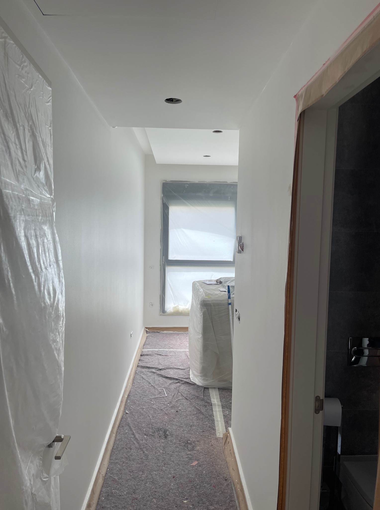 Biały korytarz w trakcie malowania: świeżo pomalowane ściany, zabezpieczone okno folią, podłoga przykryta materiałem ochronnym, widoczne wejście do łazienki.