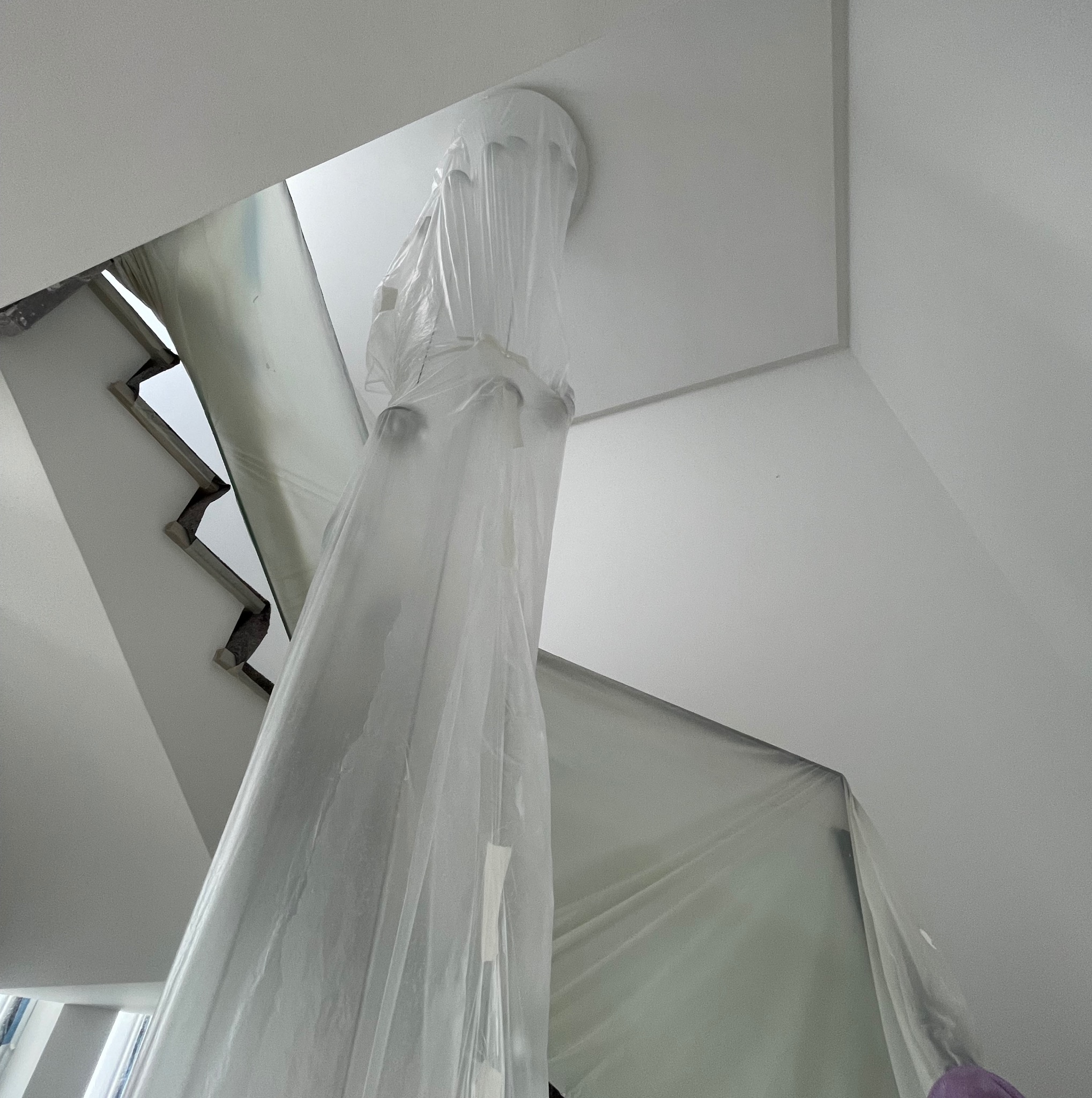 Schody i lampa zabezpieczone folią malarską w trakcie prac. Widok z dołu na białe wnętrze przed malowaniem. Zabezpieczenie przed zabrudzeniem.