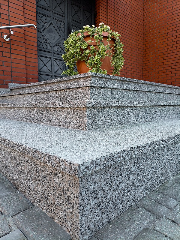 Zewnętrzne schody granitowe z donicą z kwiatami, prowadzące do ciemnych drzwi w ceglanej ścianie. Widoczny fragment chodnika z kostki brukowej.