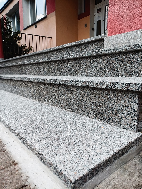 Zewnętrzne schody z szarego, nakrapianego granitu prowadzące do budynku z beżowo-czerwoną fasadą, widoczne barierki i drzwi wejściowe.