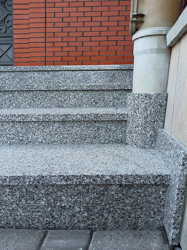 Granitowe schody zewnętrzne z zaokrąglonymi krawędziami, prowadzące do ceglanego budynku z metalową kratą w oknie, oświetlone naturalnym światłem.