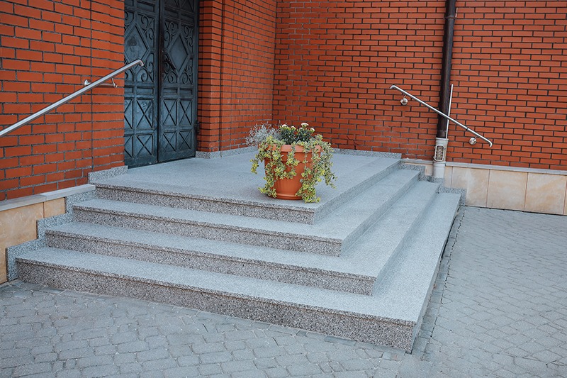 Czterostopniowe schody z szarego kamienia prowadzą do ciemnych drzwi. Na najwyższym stopniu stoi donica z kwiatami. Czerwona ceglana ściana w tle.