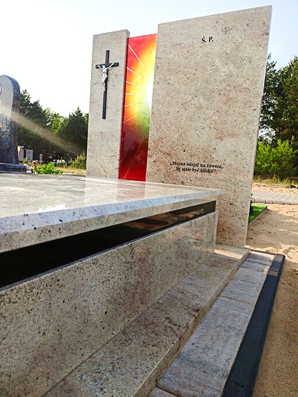 Granitowy nagrobek z krzyżem i kolorowym panelem, otoczony zielenią cmentarza. Widoczny detal kamienia i inskrypcja. Jasne, naturalne oświetlenie.