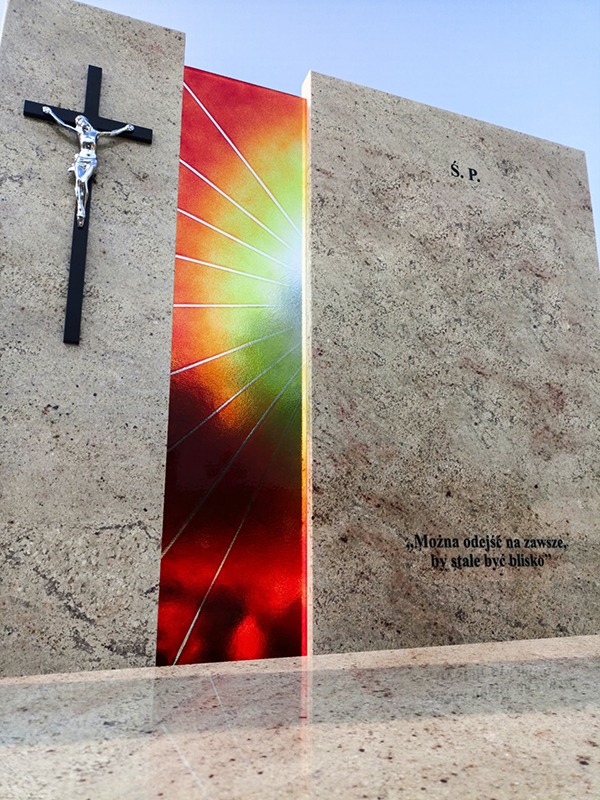 Nowoczesny, jasny nagrobek z granitu z kolorowym, podświetlonym elementem i krzyżem, z inskrypcją 'S.P.' i sentencją. Widok z dołu, podkreślający monumentalność.