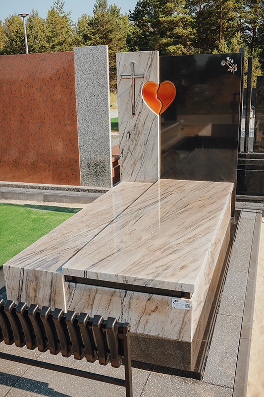 Jasny nagrobek z beżowego kamienia z motywem krzyża i serca. W tle inne nagrobki z różnych materiałów. Widoczne metalowe elementy ławki.