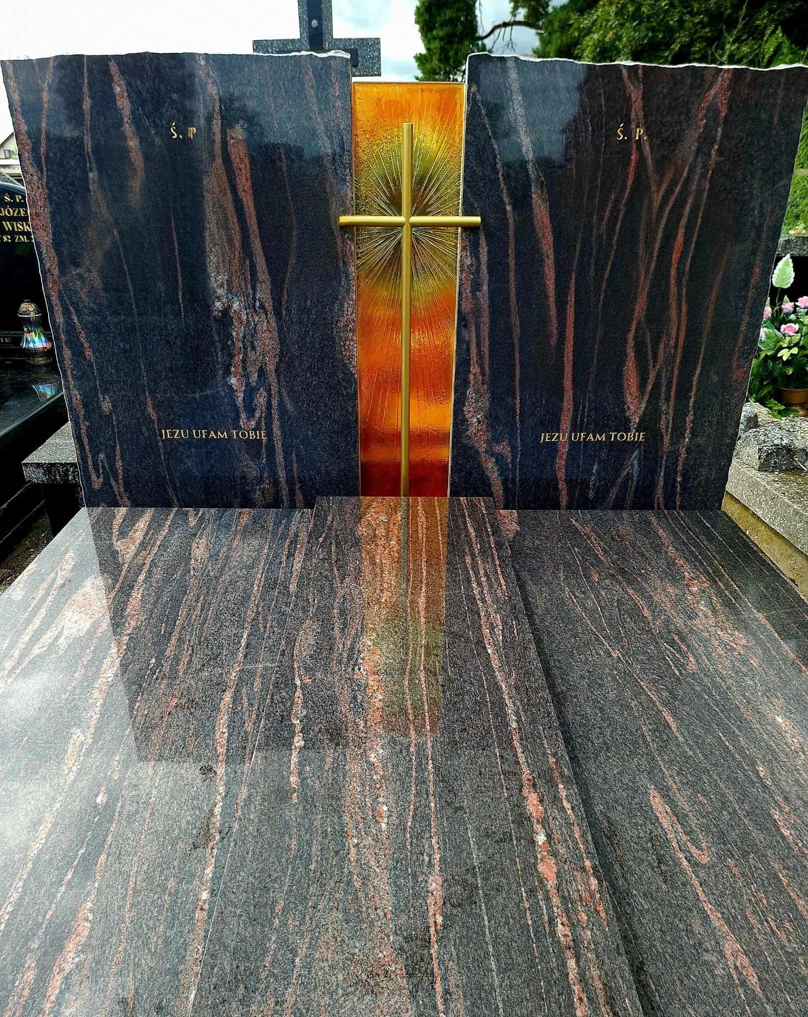 Czarny nagrobek z czerwonymi żyłkami i złotym krzyżem. Napis 'Jezu ufam Tobie'. Widoczny połysk kamienia i odbicie.