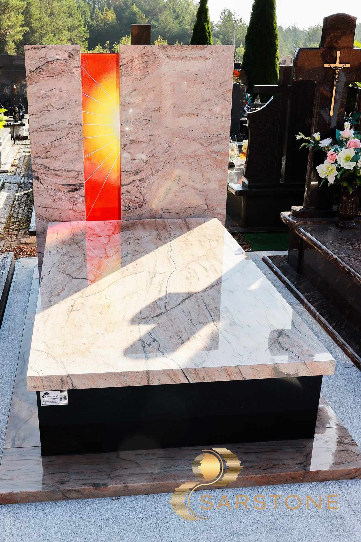 Jasnoróżowy nagrobek z motywem słońca na cmentarzu, wykonany z kamienia z widocznym użyleniem. Na podstawie naklejka z kodem QR.