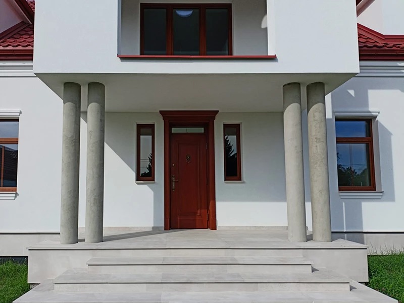 Wejście do domu z kamiennymi schodami, kolumnami i drewnianą stolarką okienną w kolorze brązu. Jasna elewacja, trzy stopnie schodów.