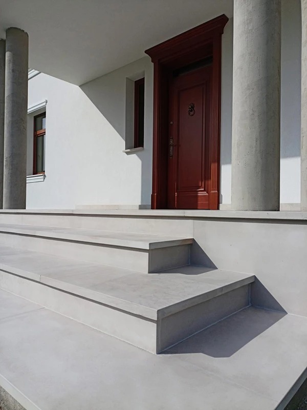 Zewnętrzne schody z jasnego kamienia prowadzące do wejścia z ciemnoczerwonymi drzwiami, wsparte kolumnami. Minimalistyczny design, naturalne światło.