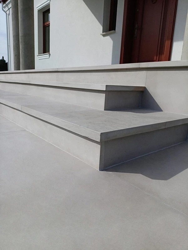 Zewnętrzne schody kamienne prowadzące do budynku z kolumnami. Minimalistyczny design, jasna kolorystyka, wyraźne cienie. Widoczne drzwi wejściowe i okno.