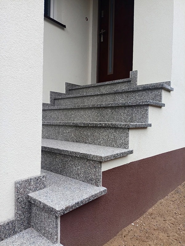 Zewnętrzne schody granitowe prowadzące do drzwi wejściowych budynku. Szare stopnie z widoczną strukturą kamienia, biała elewacja budynku.
