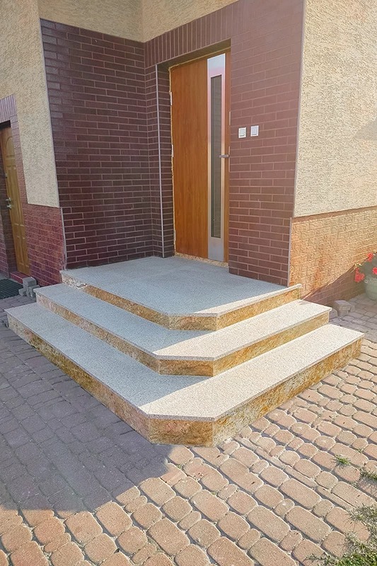 Zewnętrzne schody wejściowe z szarego kamienia kompozytowego, wykończone brązową okładziną, prowadzące do drzwi wejściowych z brązowej cegły.