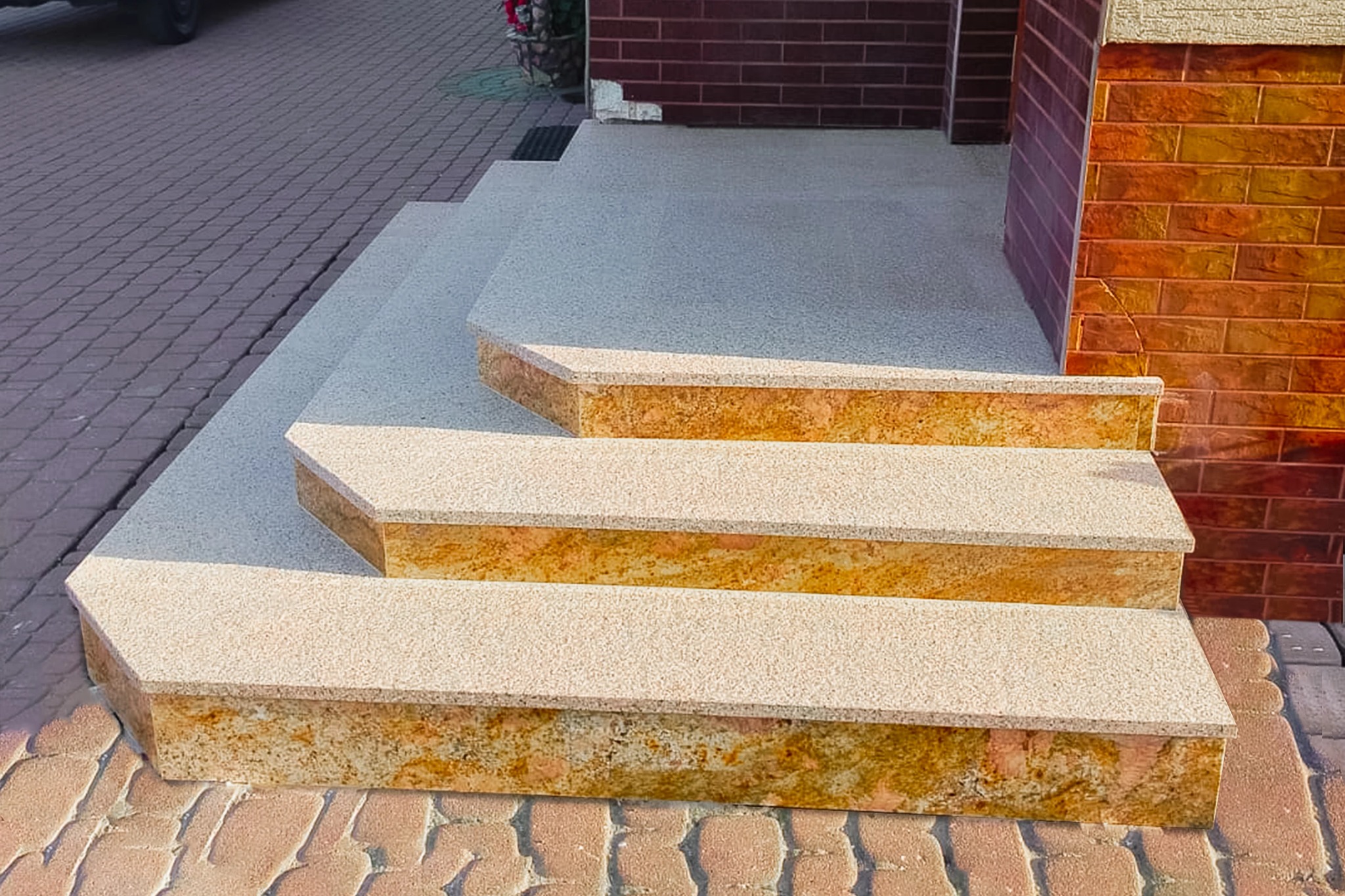 Zewnętrzne schody wejściowe z konglomeratu w odcieniach beżu i brązu, z widoczną strukturą kamienia, prowadzące do domu z ceglaną elewacją.