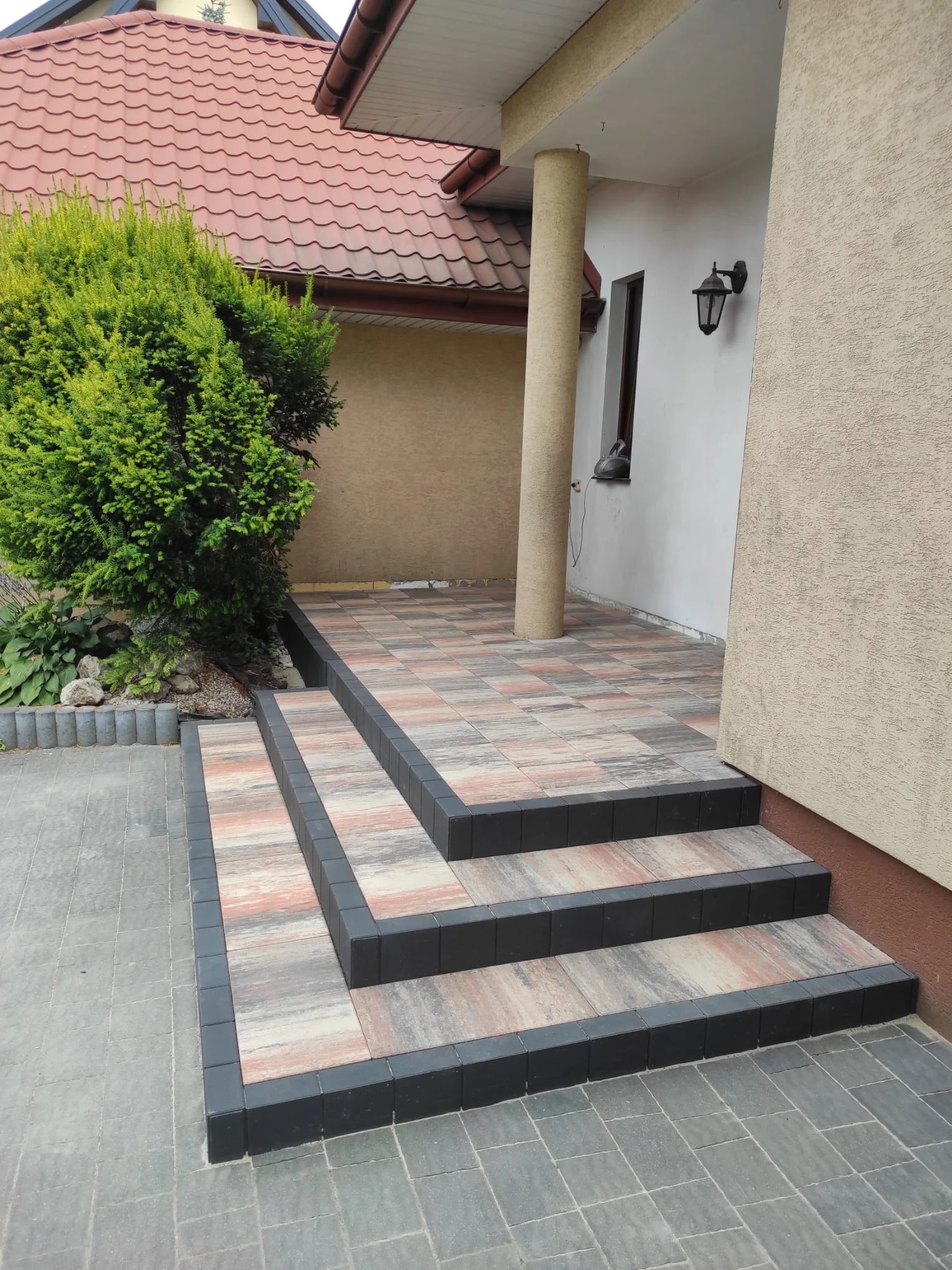 Zewnętrzne schody z kostki brukowej w odcieniach szarości, beżu i różu, obramowane czarnym klinkierem. Widok na wejście do domu z kolumną i fragmentem elewacji.