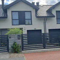 Bliźniak z ciemną stolarką okienną i grafitowym dachem za nowoczesnym, poziomym ogrodzeniem. Żółta skrzynka gazowa przy bramie. Ujęcie z poziomu chodnika.