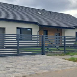 Nowoczesny dom jednorodzinny z szarą, poziomą, metalową bramą i ogrodzeniem. Na podjeździe kostka brukowa w odcieniach szarości. Widoczny fragment samochodu i kosze na śmieci.