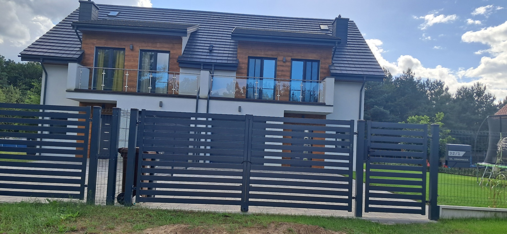 Dwukondygnacyjny dom z drewnianą elewacją i szarą, poziomą bramą wjazdową. Widoczny fragment zielonego trawnika i drzewa w tle.