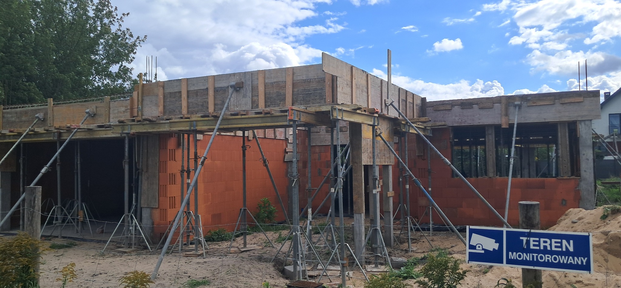 Budowa domu w trakcie: widoczne ceglane ściany, drewniane szalunki i podpory. Tablica 'Teren monitorowany' sugeruje zabezpieczenie placu budowy.