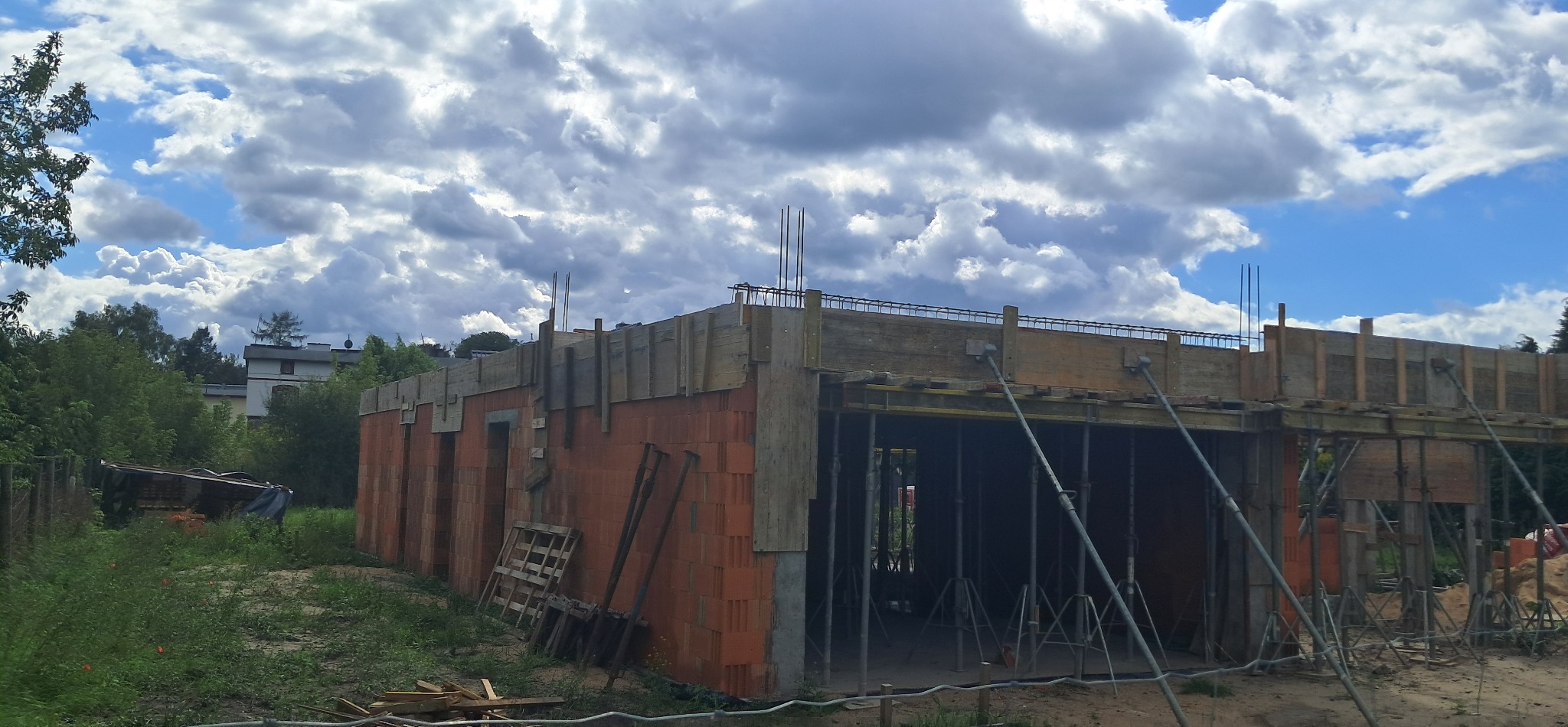Budowa domu z czerwonej cegły, widoczne zbrojenia i drewniane szalunki. W tle błękitne niebo z chmurami i zieleń drzew. Na pierwszym planie ziemia i materiały budowlane.