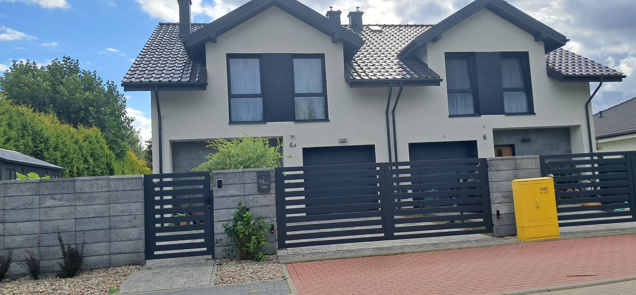 Bliźniak z ciemną stolarką okienną i grafitowym dachem za nowoczesnym, poziomym ogrodzeniem. Żółta skrzynka gazowa przy bramie. Ujęcie z poziomu chodnika.